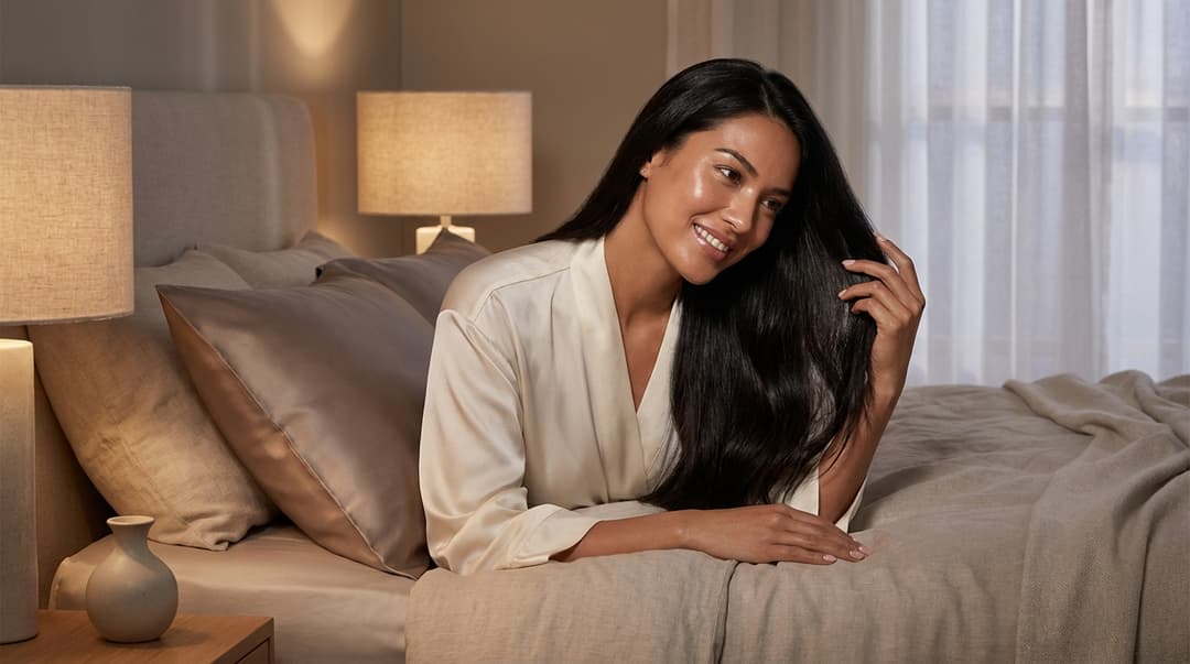Woman with glossy hair and glowing skin relaxing on a bed with a silk pillowcase
