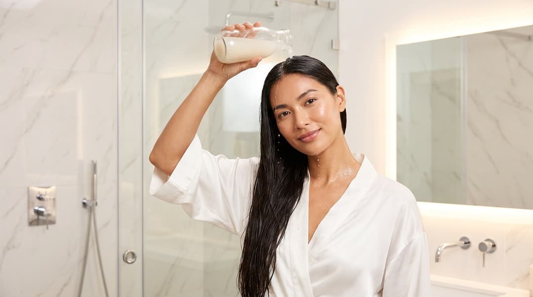 Woman with glowing skin and long hair pouring rice water over her hair in a bright modern shower