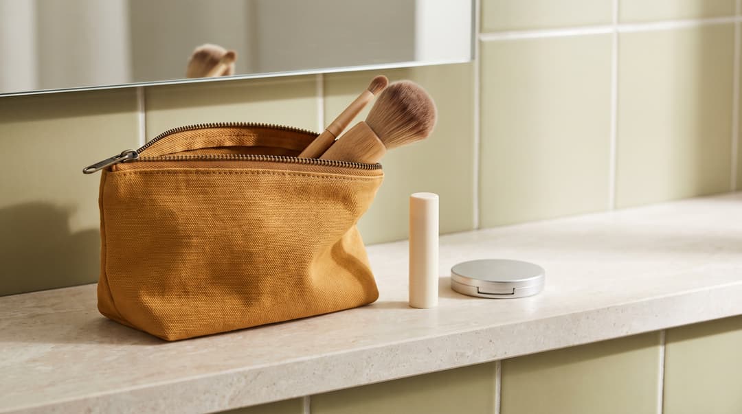 An open canvas makeup pouch with brushes, a lip balm and a compact on a shelf.