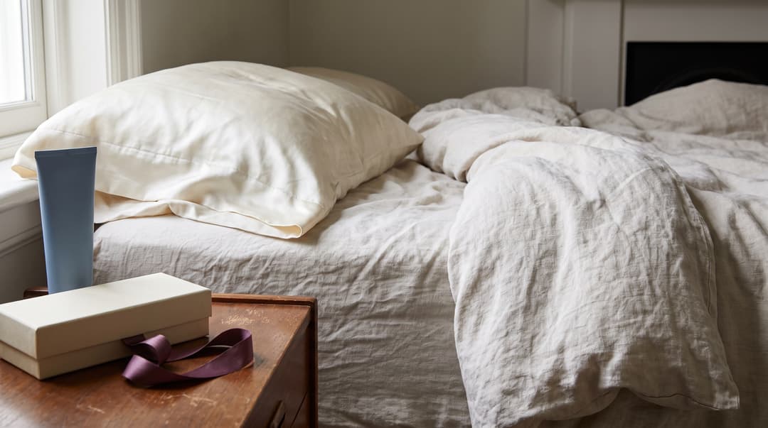 An ivory silk pillow beside a bedside table with a lotion tube and gift box.
