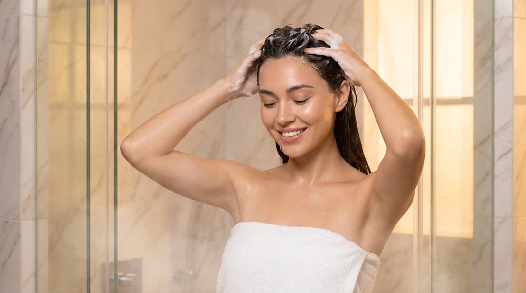 Woman gently massaging shampoo into her scalp in a marble shower.